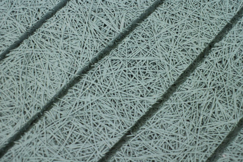 Close-up of a textured surface with overlapping, thin, light blue fibers arranged in a random pattern. Four diagonal grooves separate the fibrous areas, creating distinct sections.