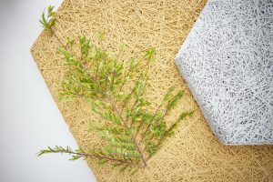 A green leafy branch lies on a tan woven mat, next to a white hexagonal woven mat, all arranged on a white background.