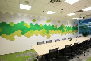 A modern conference room with a long wooden table, black office chairs, and a wall decorated with hexagonal panels in shades of green and gray. Large windows and ceiling lights brighten the space.