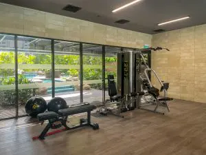 A modern gym room with a weight bench, exercise balls, and a multi-functional strength training machine, with large windows overlooking a pool and lush greenery outside.