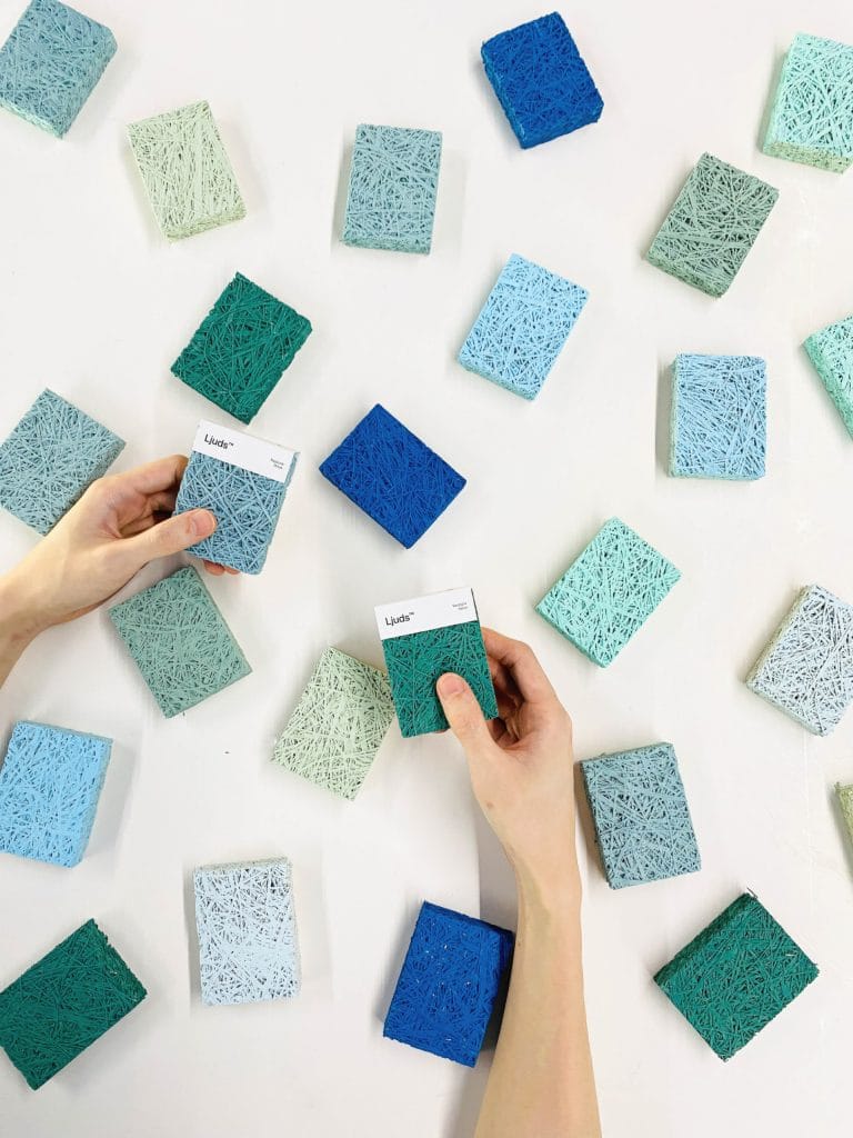 Hands holding two square textured color swatches among many similar swatches scattered on a white surface in shades of blue, green, and gray.