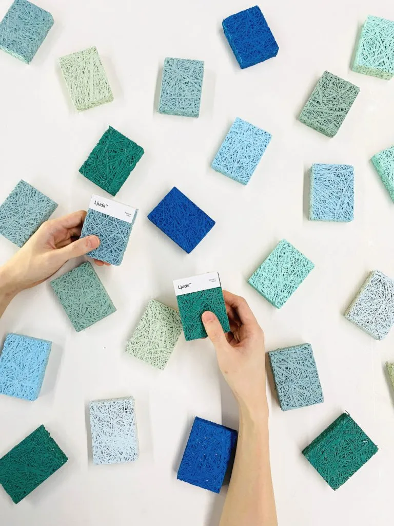 Hands holding two square textured color swatches among many similar swatches scattered on a white surface in shades of blue, green, and gray.