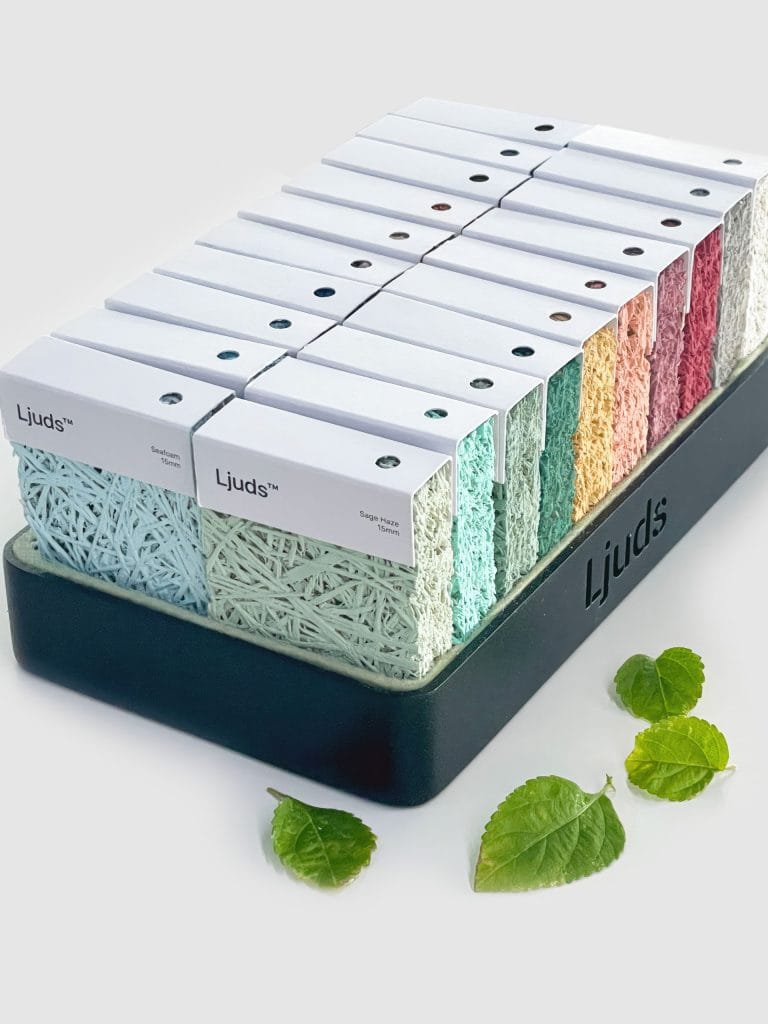 A display tray holds colorful Ljuds branded sound-absorbing panels with white tops, arranged in neat rows. Three green leaves are scattered nearby on a white surface.