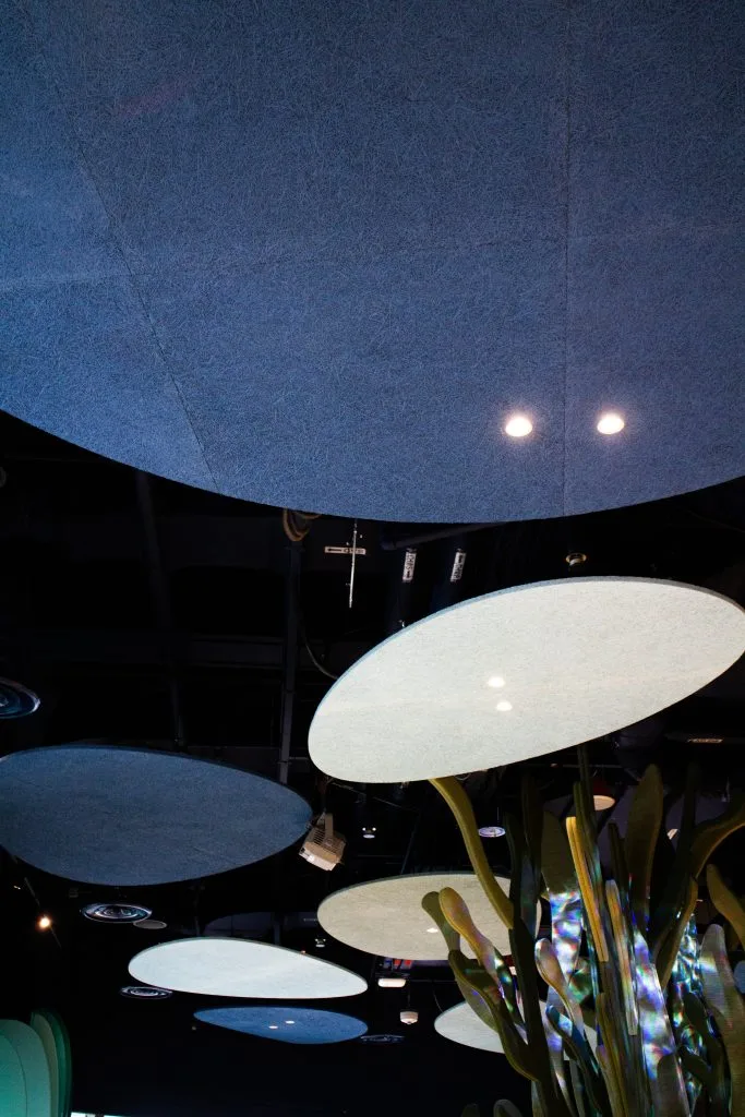 Large, round, blue and white panels hang from a dark ceiling. Two ceiling lights shine, and green plant-like structures are visible at the bottom right. The setting appears modern and artistic.