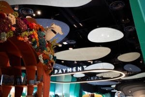 A playful indoor space with colorful, coral-like decorations, a white shark model, shelves of books, and a large PAYMENT sign hanging from the ceiling with circular and oval ceiling panels above.