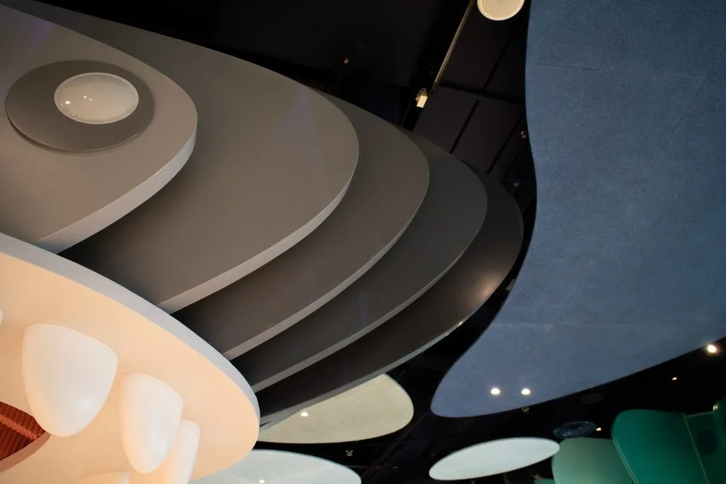 Curved, layered gray and blue ceiling panels with smooth, abstract shapes and built-in circular lights, viewed from below in a modern indoor space.