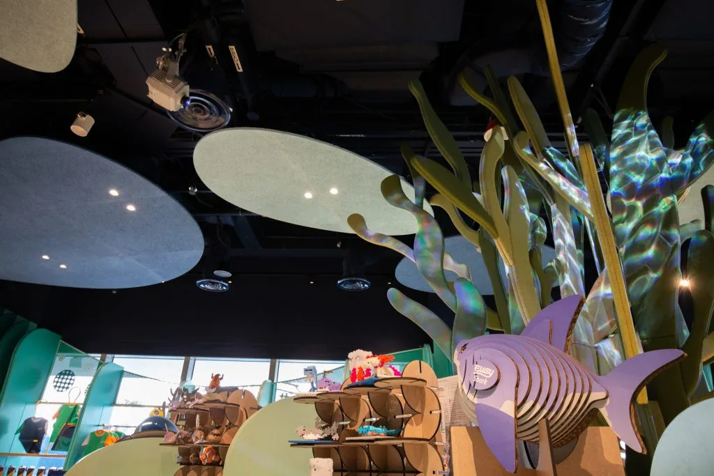 A playful indoor scene features green seaweed-like structures, large round ceiling panels, and a purple cardboard fish decoration above shelves displaying various toys and animal figurines. Natural light streams in from windows.