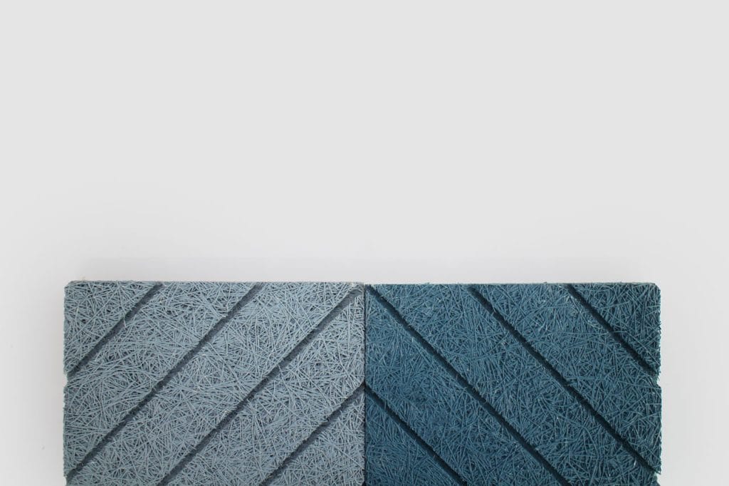 Two rectangular panels with diagonal grooves, one light gray and one dark blue, are placed side by side against a plain white background. Both panels have a textured, fibrous surface.