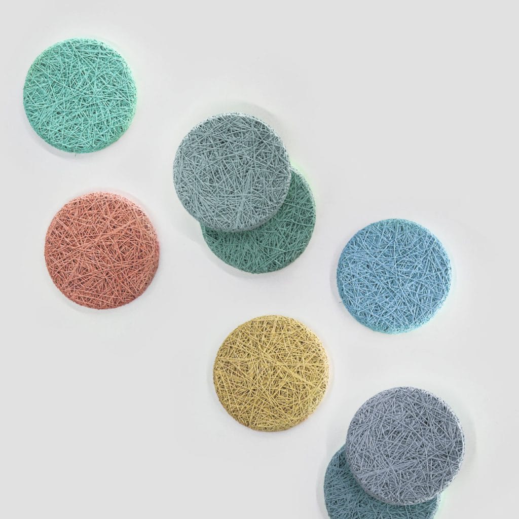Eight round, textured coasters in pastel colors (teal, coral, blue, yellow, and gray) are arranged on a light gray surface in a scattered pattern.