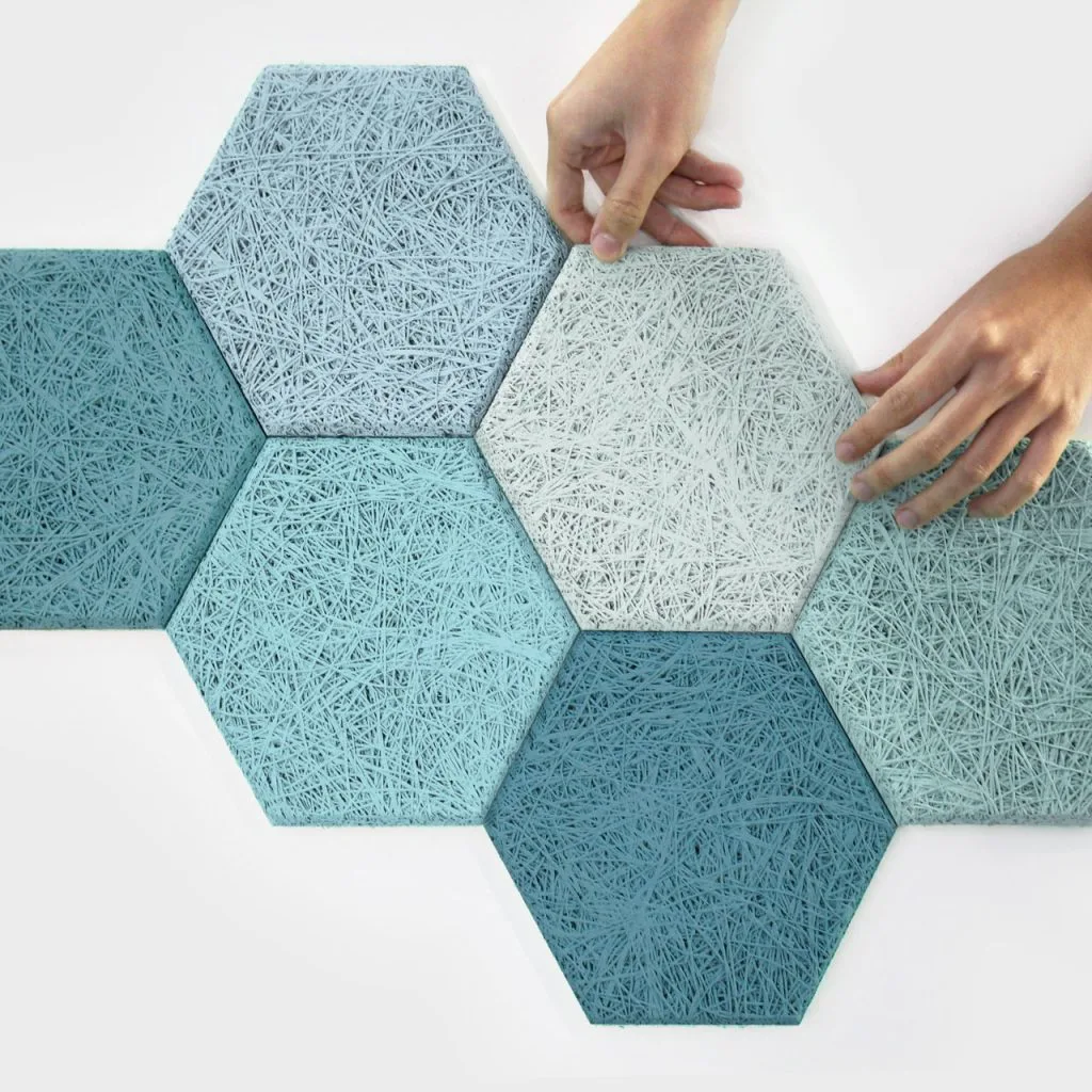 A pair of hands arranges textured hexagonal tiles in shades of blue and white on a white surface. The tiles have a woven, fibrous appearance and fit together in a honeycomb pattern.