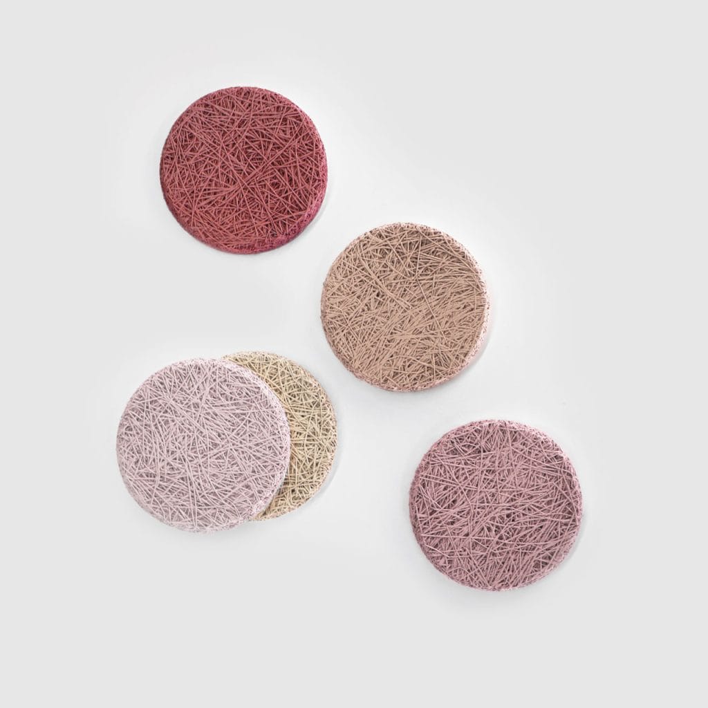 Five round, textured coasters in muted shades of red, pink, beige, and lavender are arranged on a light gray background. The coasters feature a tangled, string-like surface pattern.