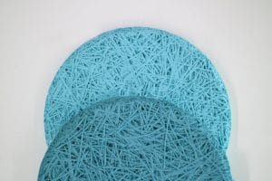 Two round, textured mats in light and dark blue overlap each other on a white background. The mats have a woven, string-like pattern, creating a layered and intricate design.