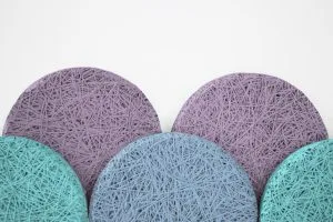 Five circular coasters, three purple and two teal, feature a tangled string pattern on their surfaces and are arranged overlapping each other against a white background.