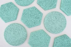 Several teal-colored coasters in round and hexagonal shapes, featuring a textured, woven string pattern, are arranged on a white surface.