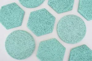 Several teal-colored coasters in round and hexagonal shapes, featuring a textured, woven string pattern, are arranged on a white surface.