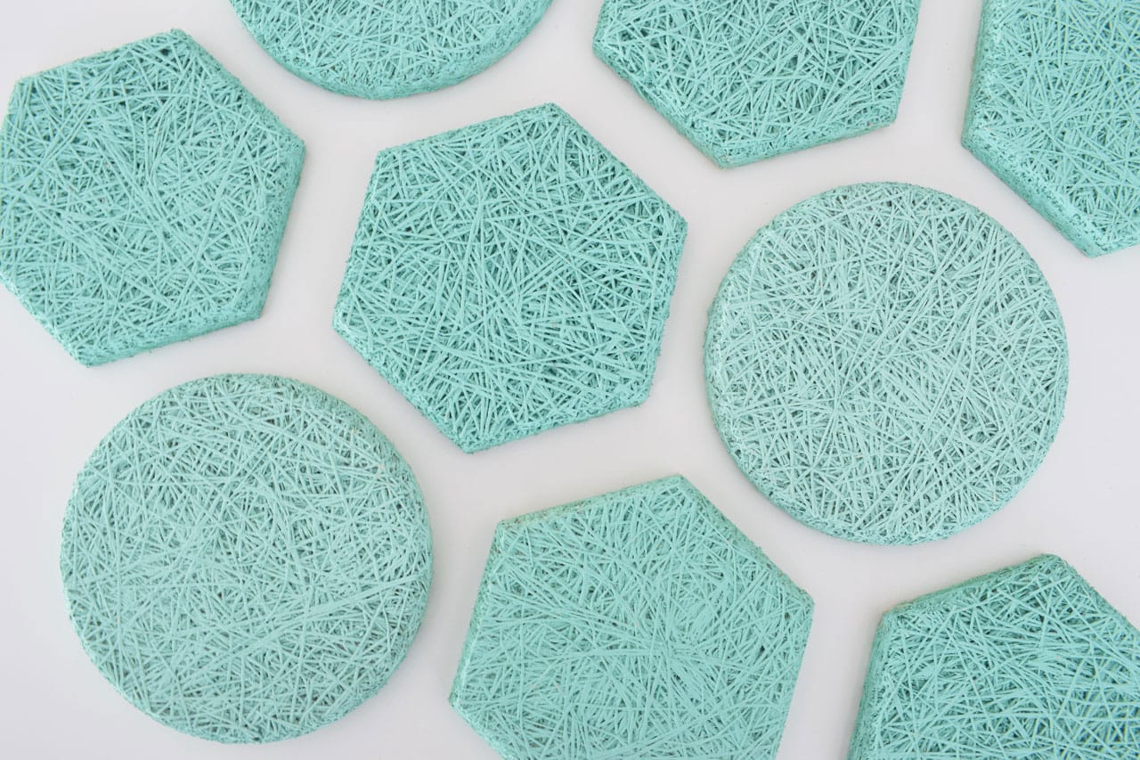 Several teal-colored coasters in round and hexagonal shapes, featuring a textured, woven string pattern, are arranged on a white surface.