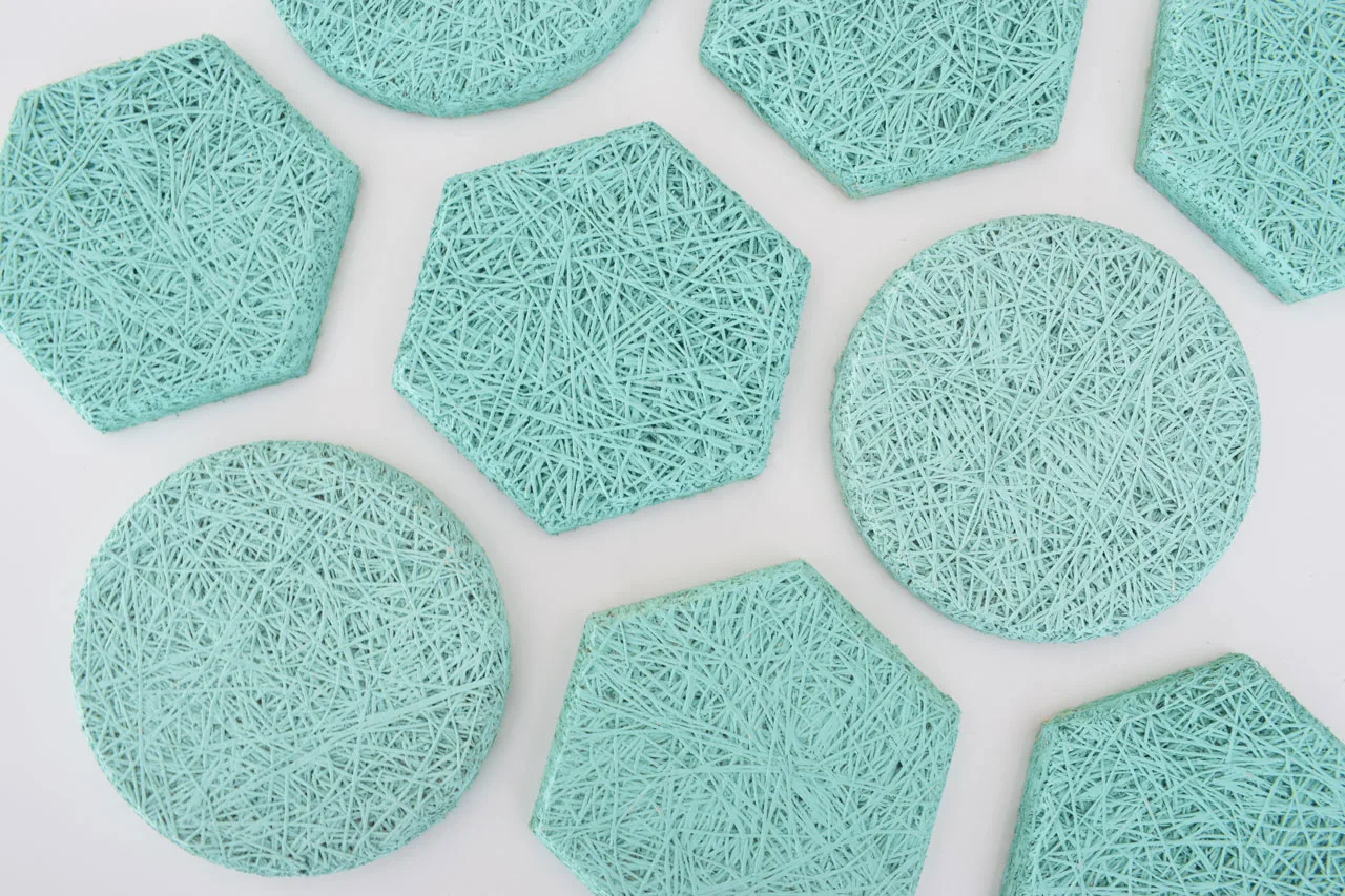 Several teal-colored coasters in round and hexagonal shapes, featuring a textured, woven string pattern, are arranged on a white surface.