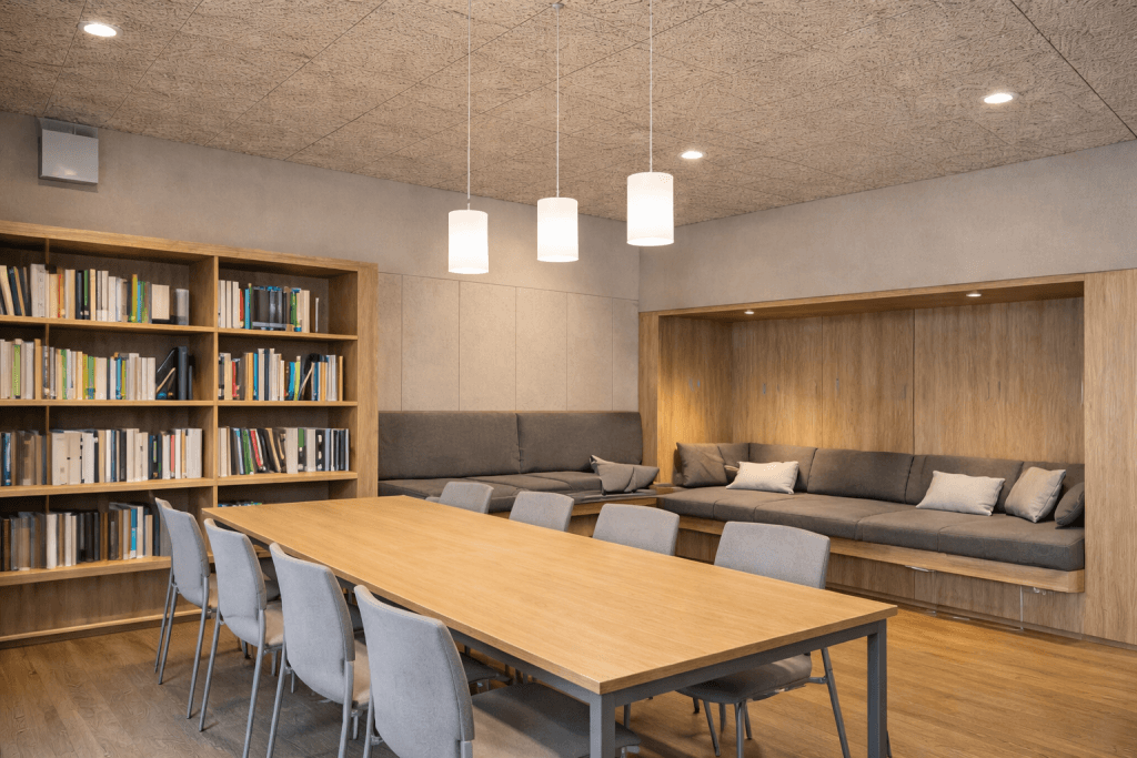 Modern study room with a long wooden table and grey chairs, built-in bookshelves filled with books, and a cozy alcove seating area with cushions under soft pendant lighting.