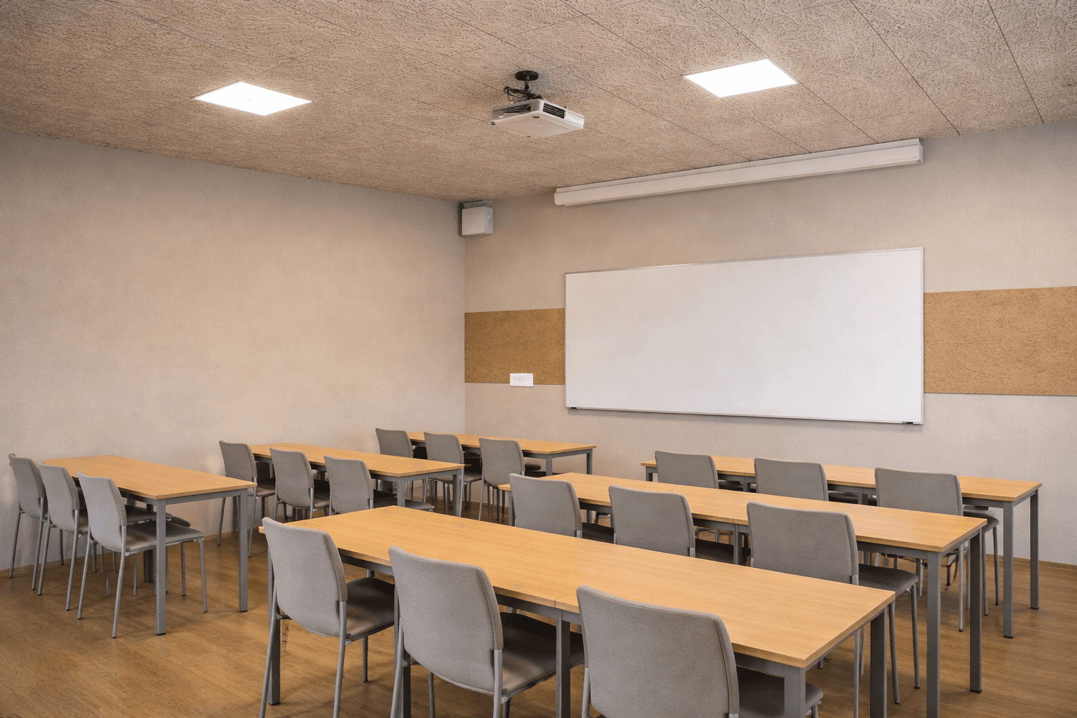A modern, empty classroom with light-colored walls, wooden desks arranged in rows, gray chairs, a large whiteboard, cork bulletin boards, and a ceiling-mounted projector.
