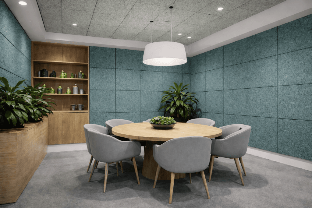 A modern meeting room with a round wooden table, six gray chairs, green indoor plants, and blue textured walls. A large white ceiling light hangs above the table, and shelves with decor are visible in the background.