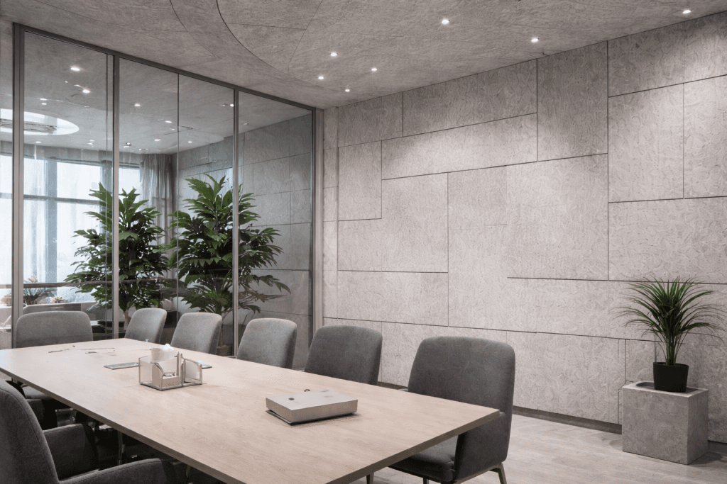 Modern conference room with a light wooden table, gray chairs, minimalist décor, large indoor plants, and textured gray walls. Glass panels separate the room from another area, allowing natural light to enter.