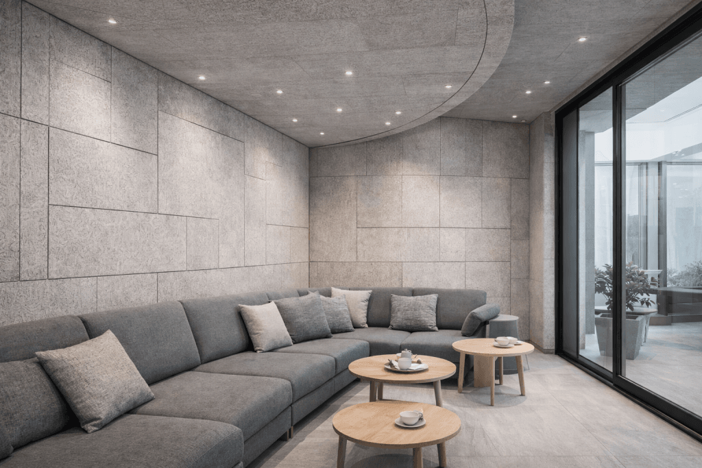 Modern lounge with gray sectional sofa, matching cushions, and two round wooden coffee tables set with cups and plates. Room features textured gray walls, recessed ceiling lights, and large glass doors overlooking a patio.