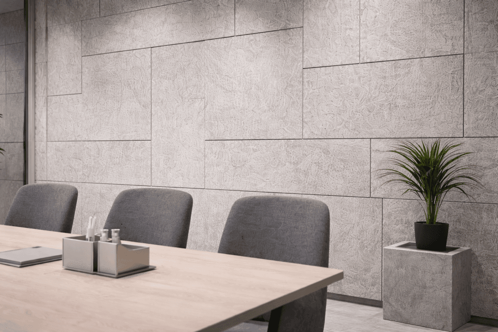 A modern meeting room with a light wooden table, gray upholstered chairs, a small metal organizer on the table, and a green potted plant on a square stand against a textured gray wall.
