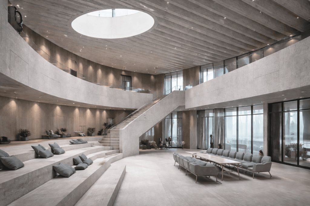 A spacious, modern interior with curved concrete walls, a central staircase, a circular skylight, tiered seating with gray cushions, lounge chairs, and floor-to-ceiling windows letting in natural light.