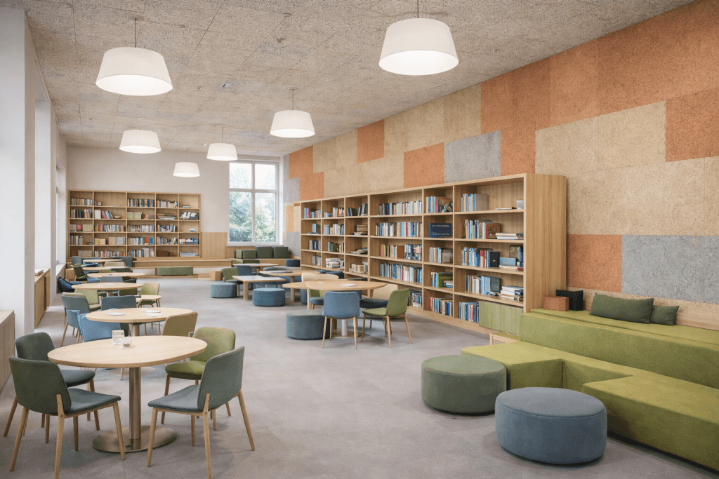 A spacious, modern library with round tables, green and blue chairs, built-in bookshelves, large windows, and cushioned seating along the wall with geometric-patterned paneling. The room is well-lit and inviting.